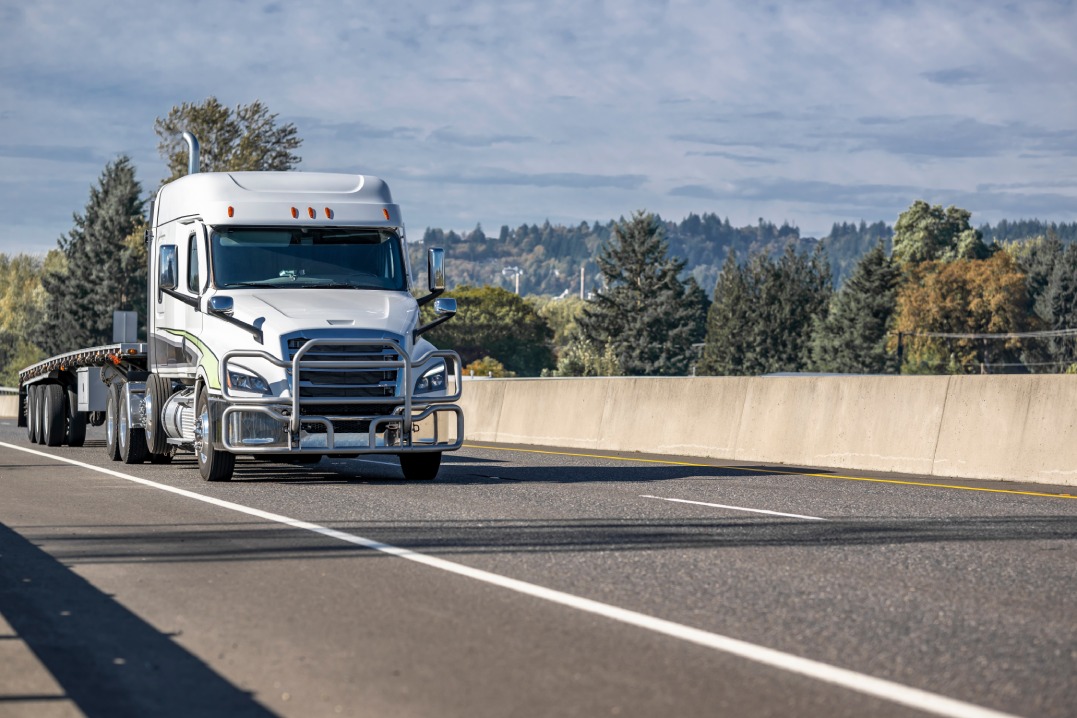 White low cab profile carrier big rig semi truck transporting flat bed semi trailer running on the one way overpass highway road with yellow autumn trees on the side going to distribution warehouse