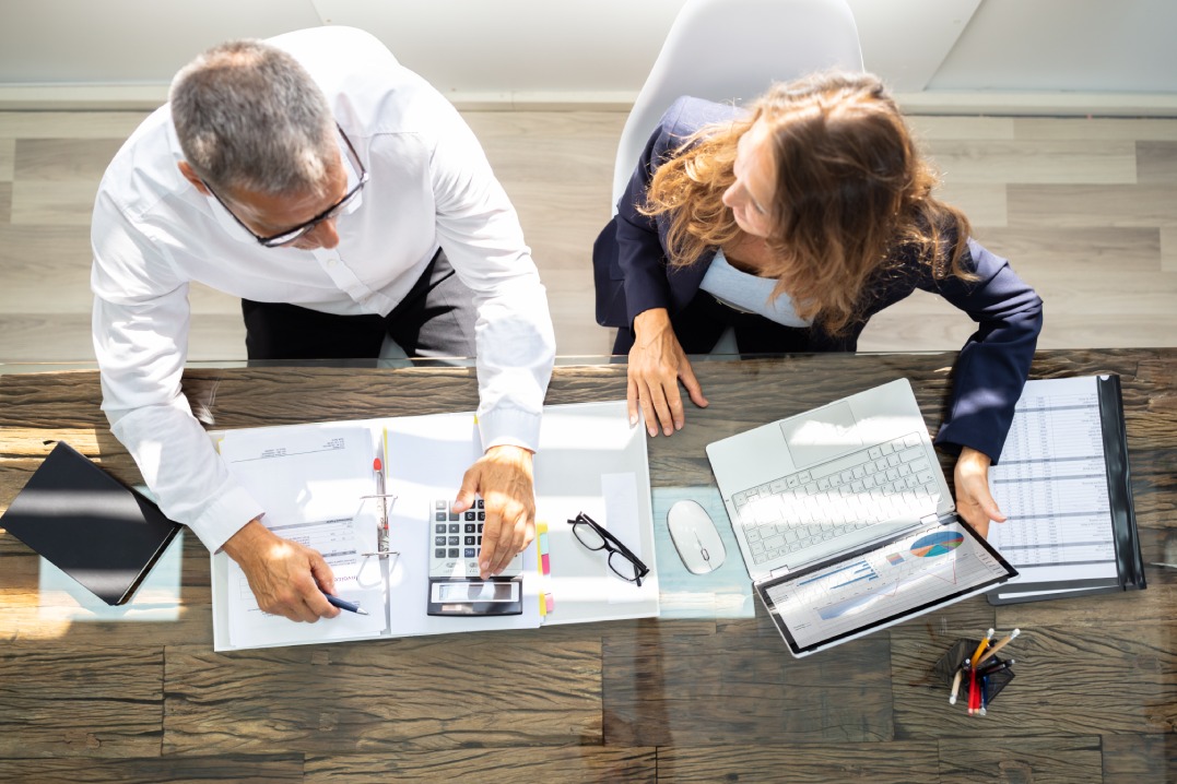 An Overhead View Of Two Businesspeople Calculating Invoice In Office While Using Laptop